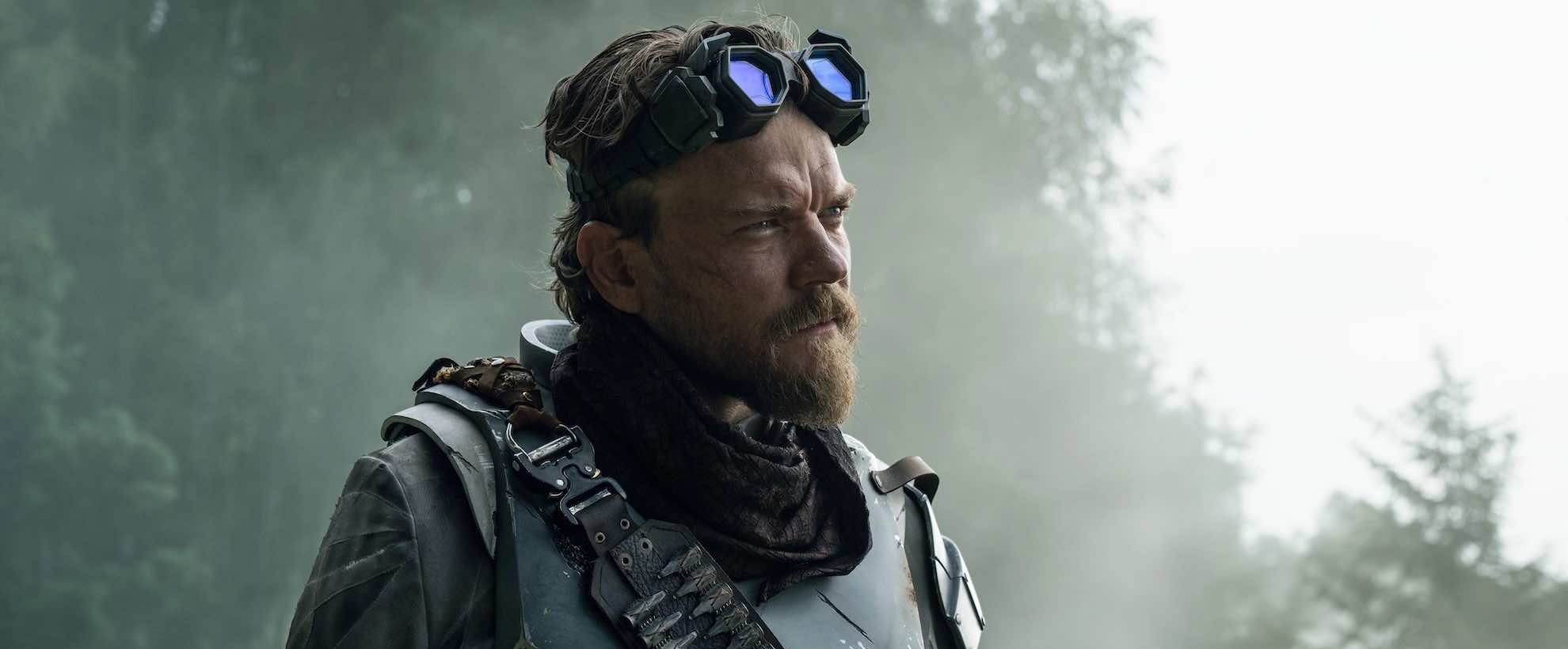 A man in post-apocalyptic fashion stands in a misty woodland